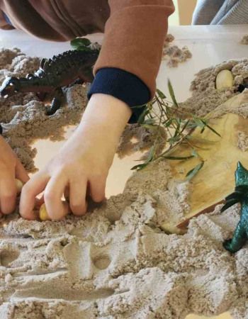 Un petit garçon jouant dans une boîte rempli de sable avec des jouets en forme de dinosaure 