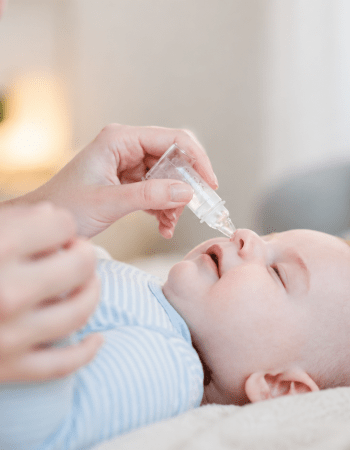 Un adulte tient délicatement un flacon de sérum physiologique au-dessus du nez d’un bébé allongé sur une couverture, dans un intérieur doux et lumineux.