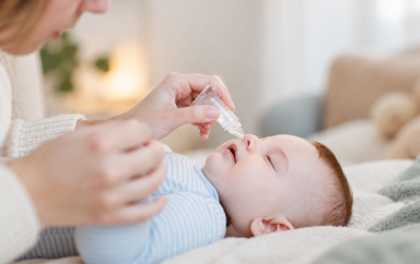 Un adulte tient délicatement un flacon de sérum physiologique au-dessus du nez d’un bébé allongé sur une couverture, dans un intérieur doux et lumineux.