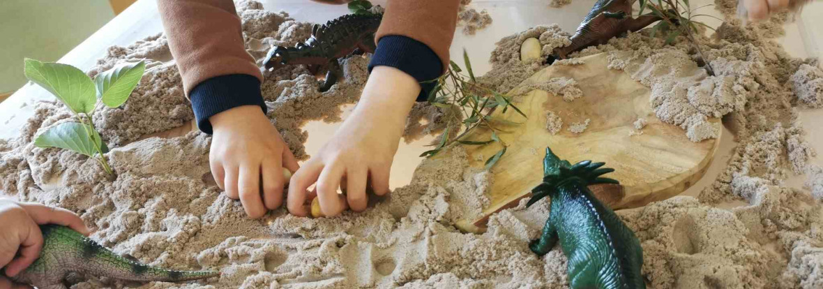 Un petit garçon jouant dans une boîte rempli de sable avec des jouets en forme de dinosaure 